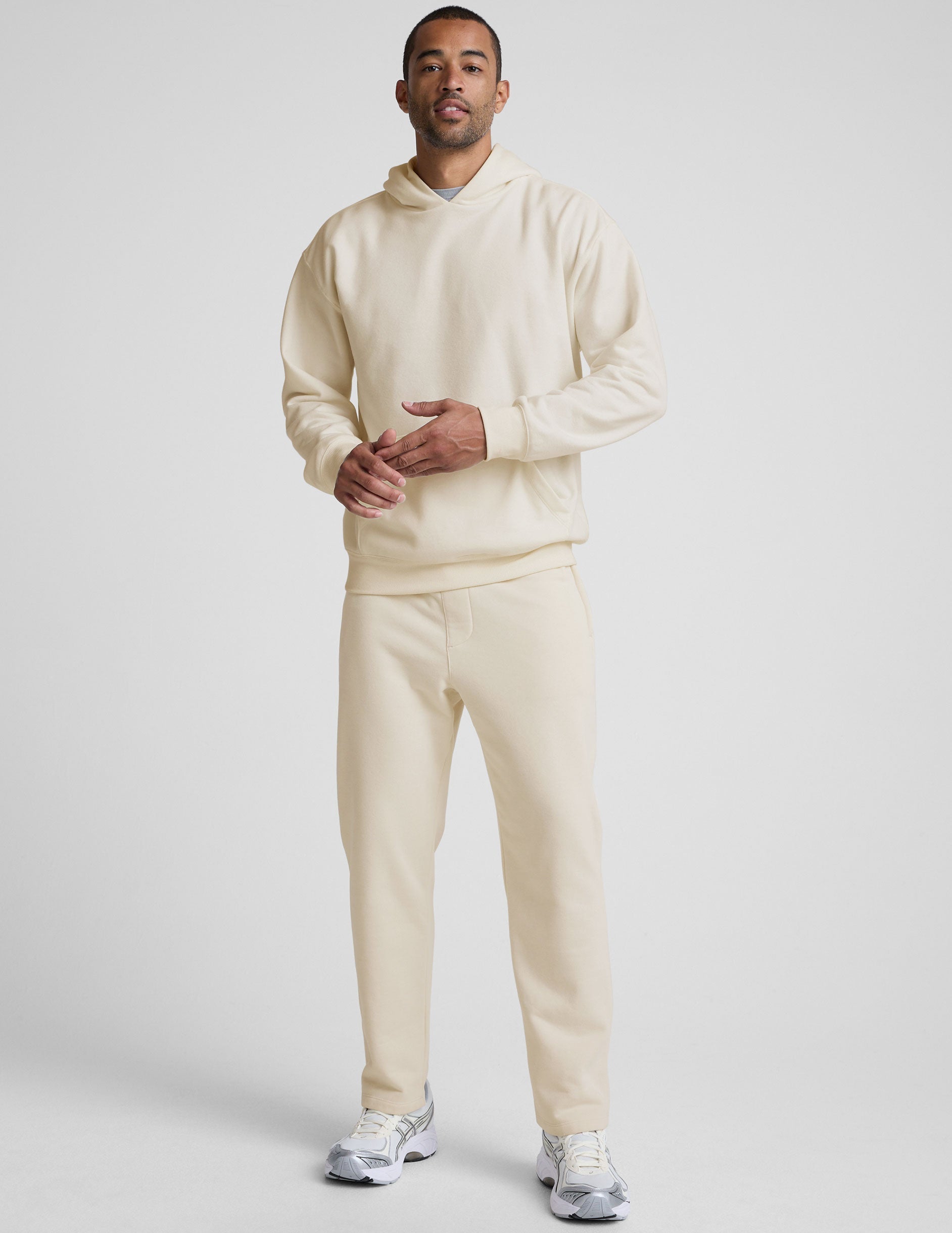 man wearing a cream-colored hoodie and straight leg fleece sweatpants on a plain background.