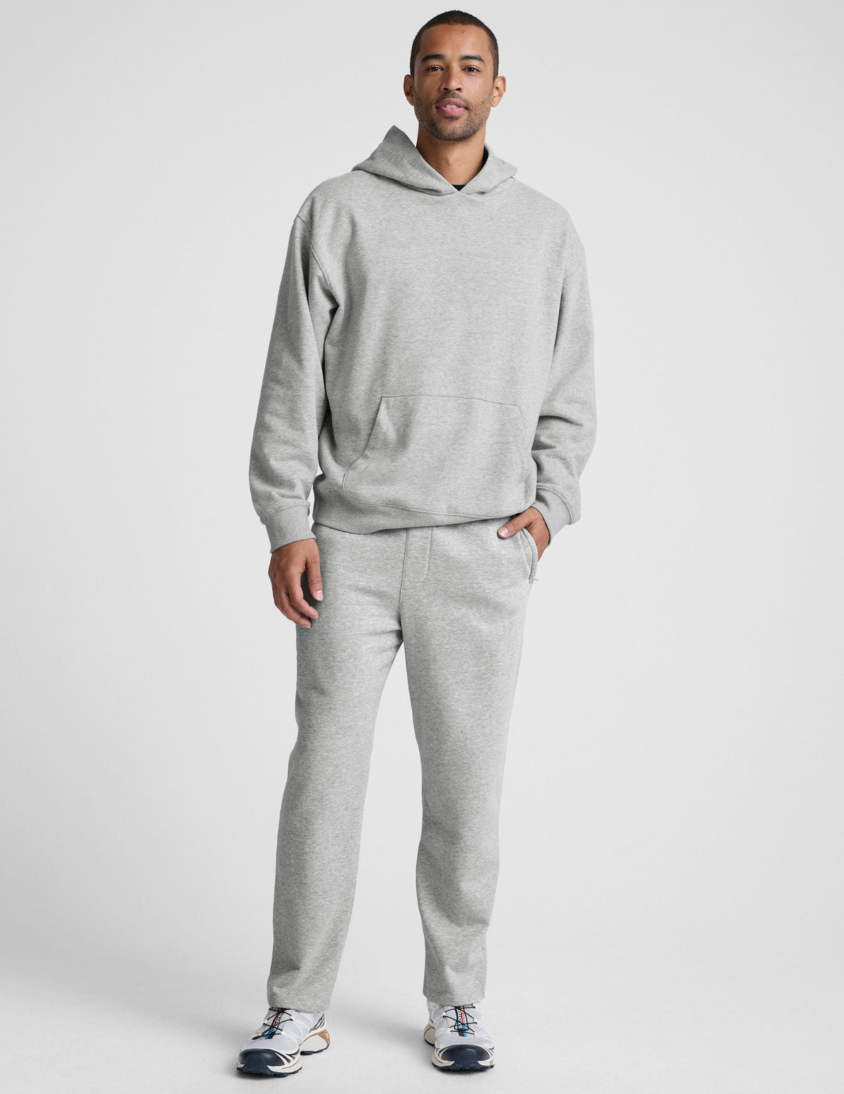 man wearing a gray hoodie and straight leg fleece sweatpants on a plain background.