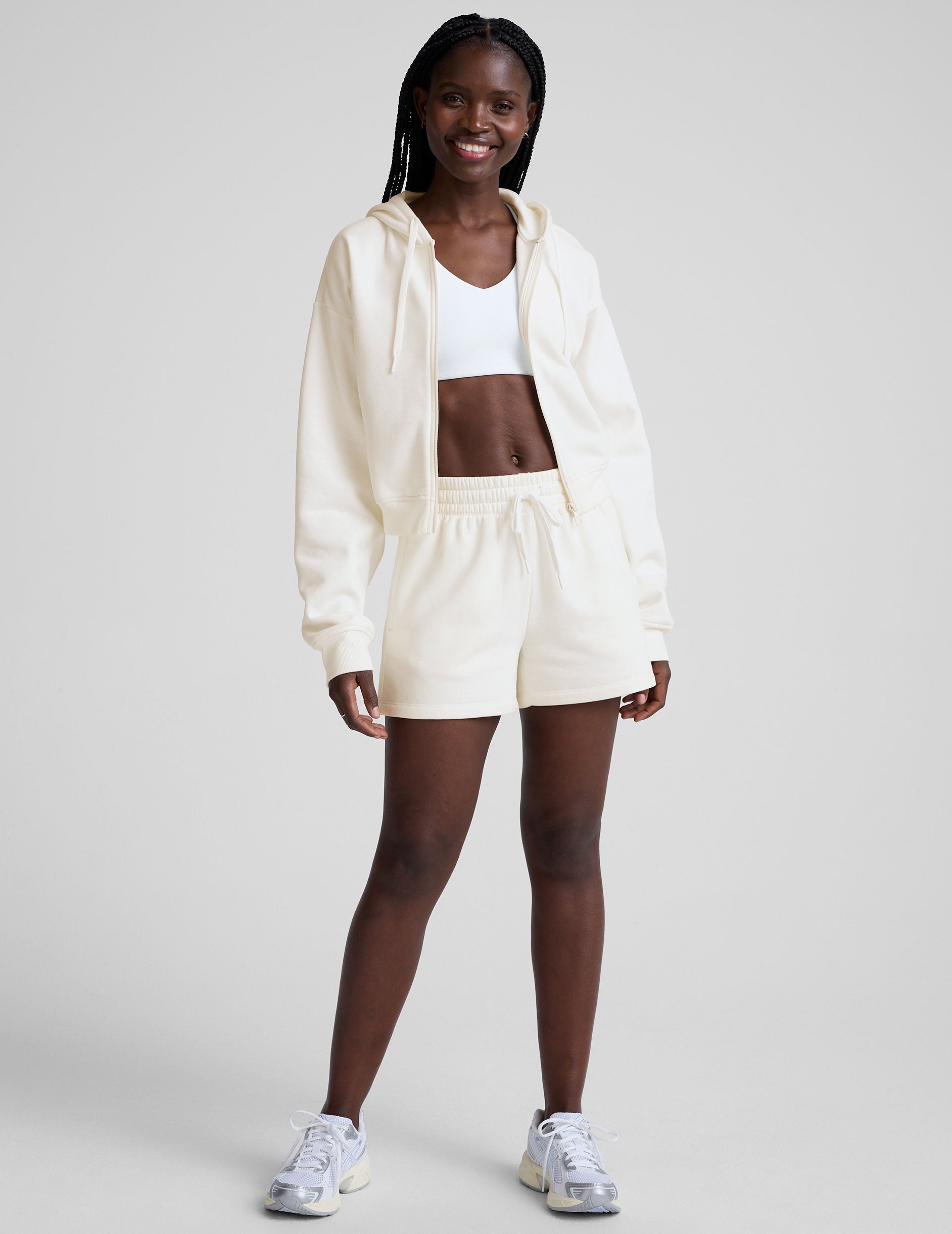 model wearing a white fleece zip-up hoodie and fleece sweat shorts with a drawcord at waistband on a plain background.