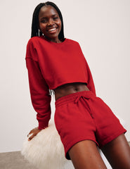 woman wearing a red pullover sweatshirt and sweat shorts against a plain background.