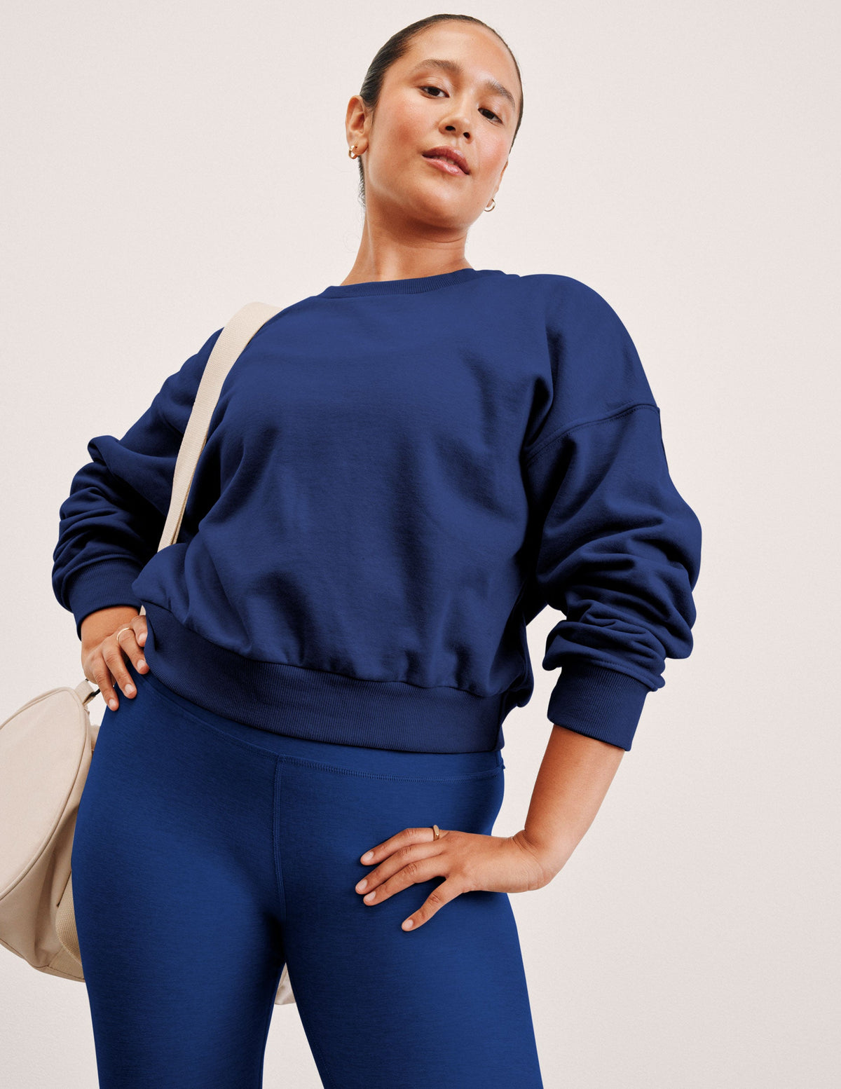 woman wearing a blue sweatshirt and leggings with a beige bag on a plain background.