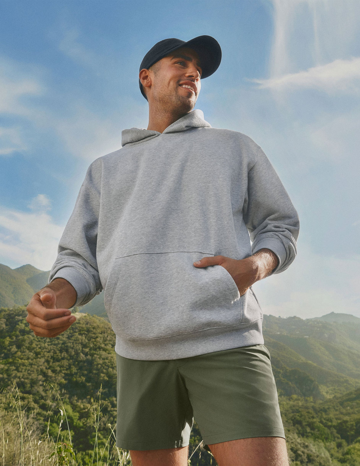 man wearing a gray hoodie and green shorts standing outdoors with mountains in the background.