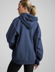 woman wearing a blue hoodie and black sweats against a plain background.