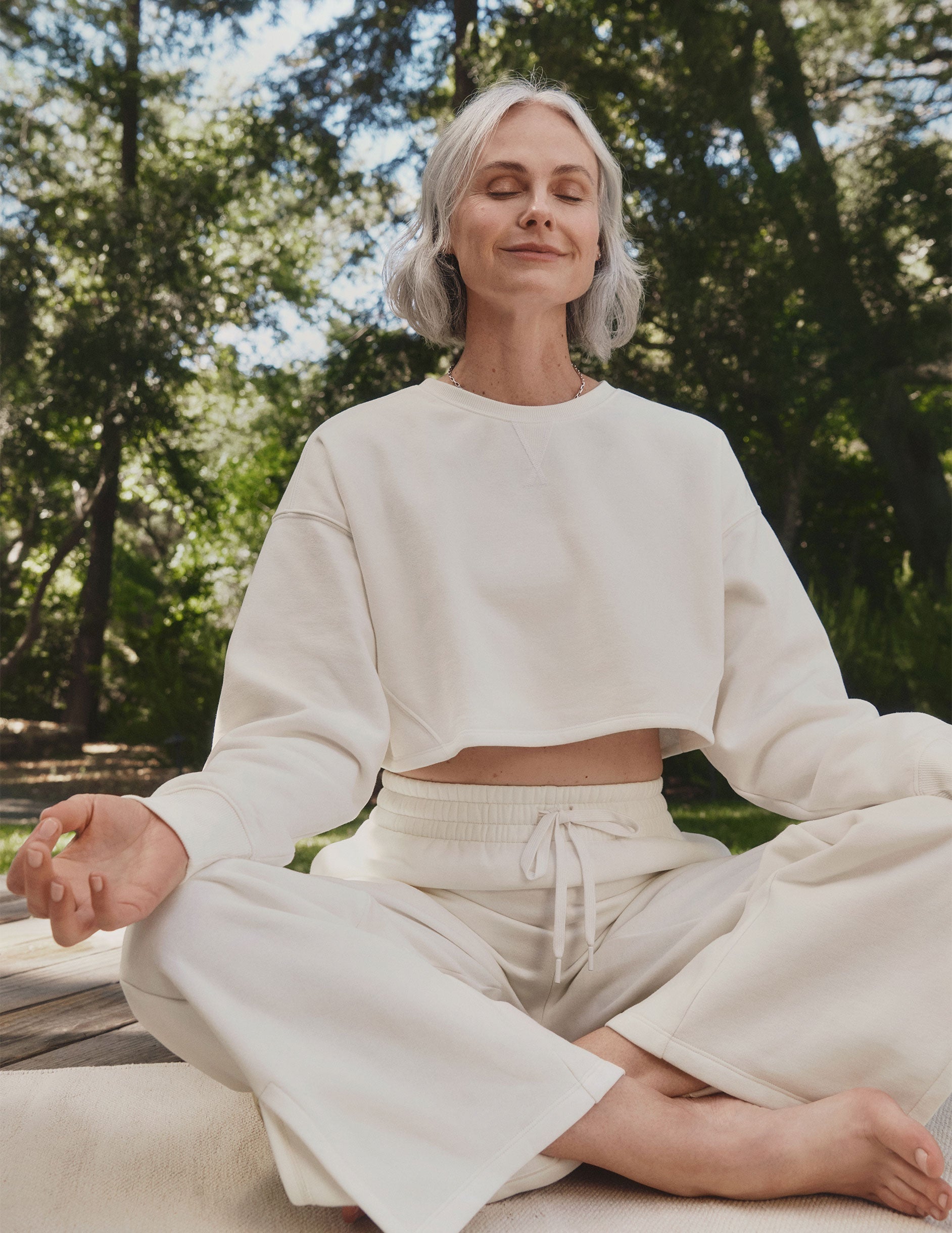 model in white boxy cropped sweatshirt and fleece wide leg pants meditating outdoors with trees in the background.