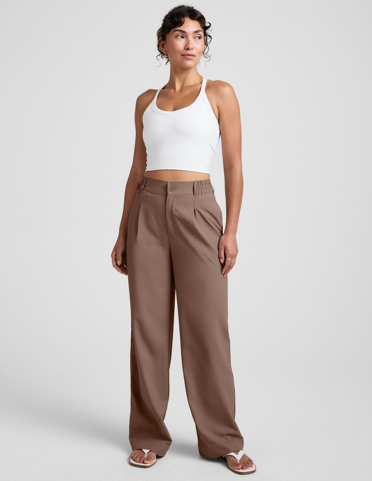 Woman wearing a white tank top and brown high-waisted trousers on a light gray background