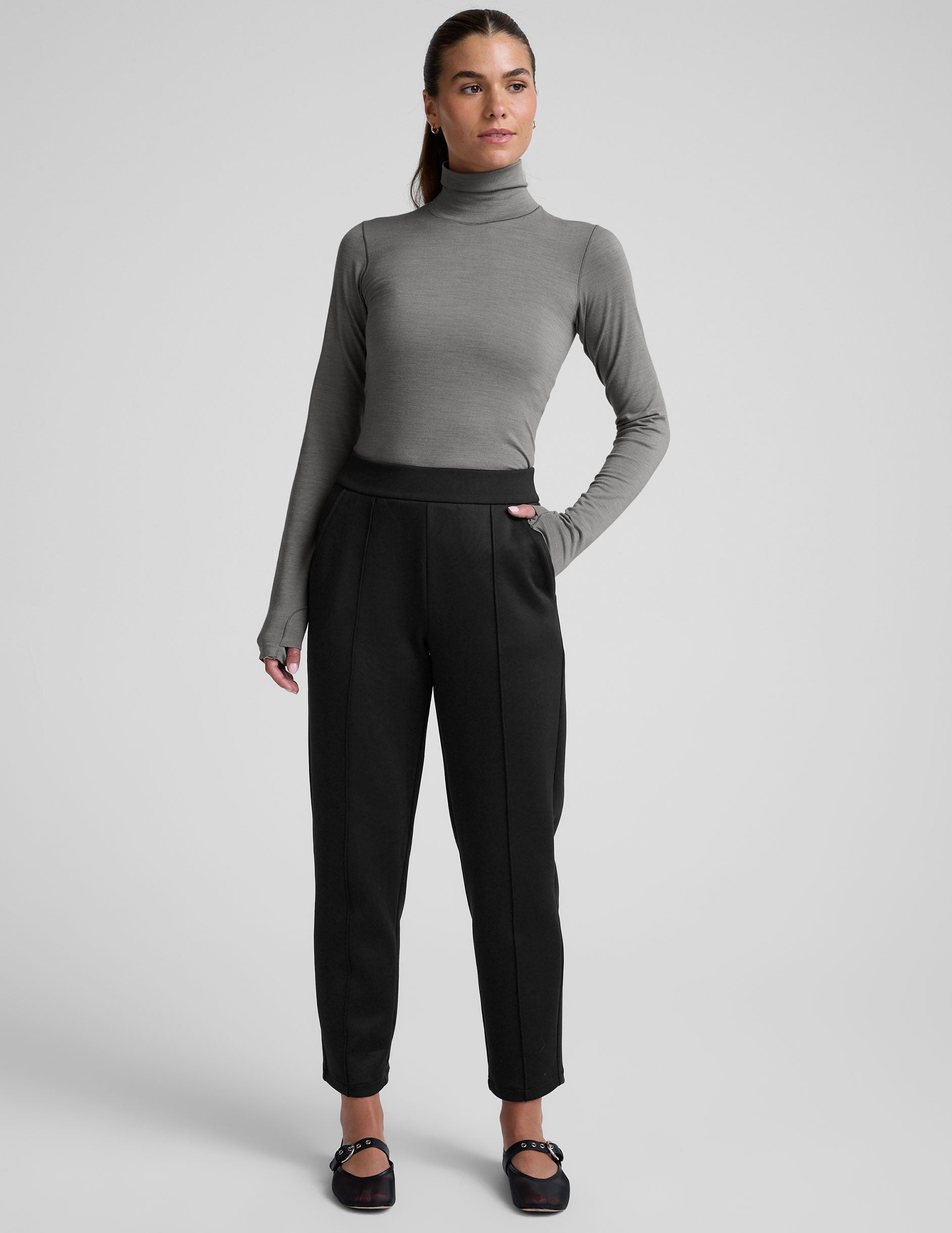 woman wearing a gray turtleneck and black tapered pants on a white background.