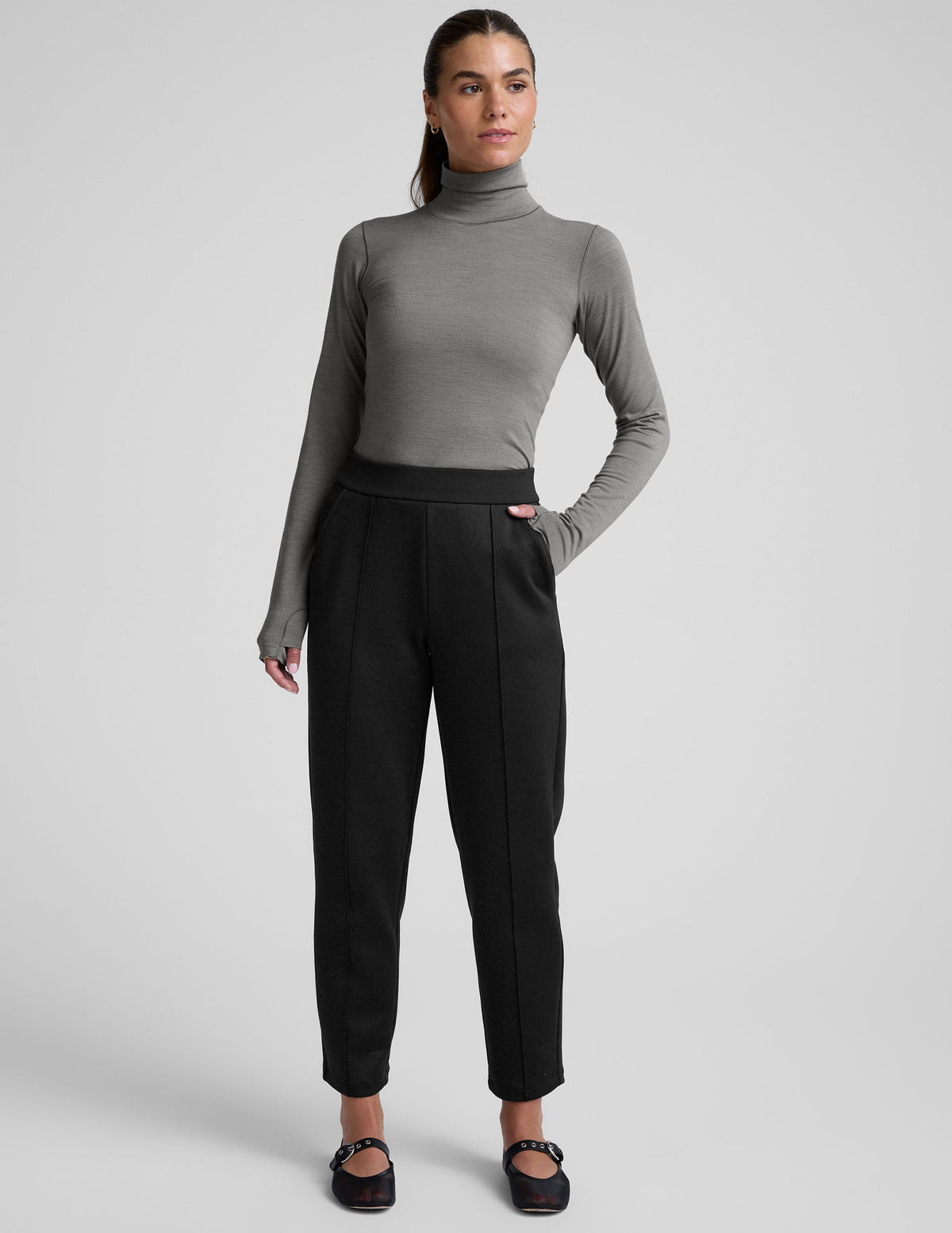 woman wearing a gray turtleneck and black tapered pants on a white background.