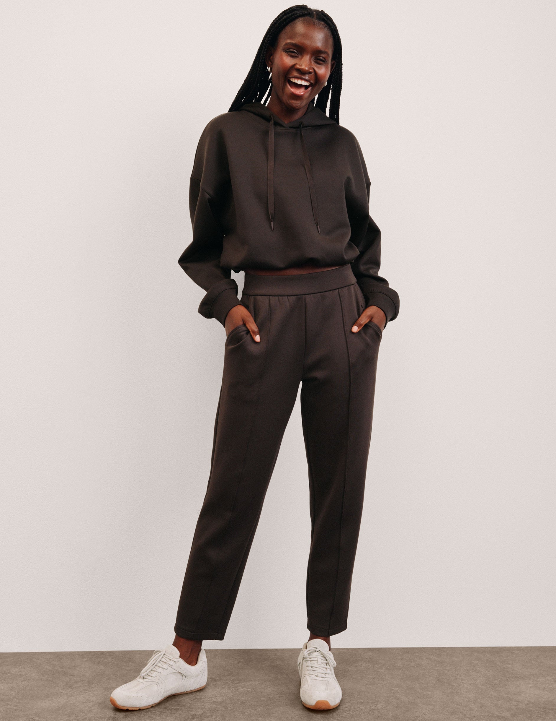 woman wearing a brown cropped hoodie and brown tapered pants against a plain background.