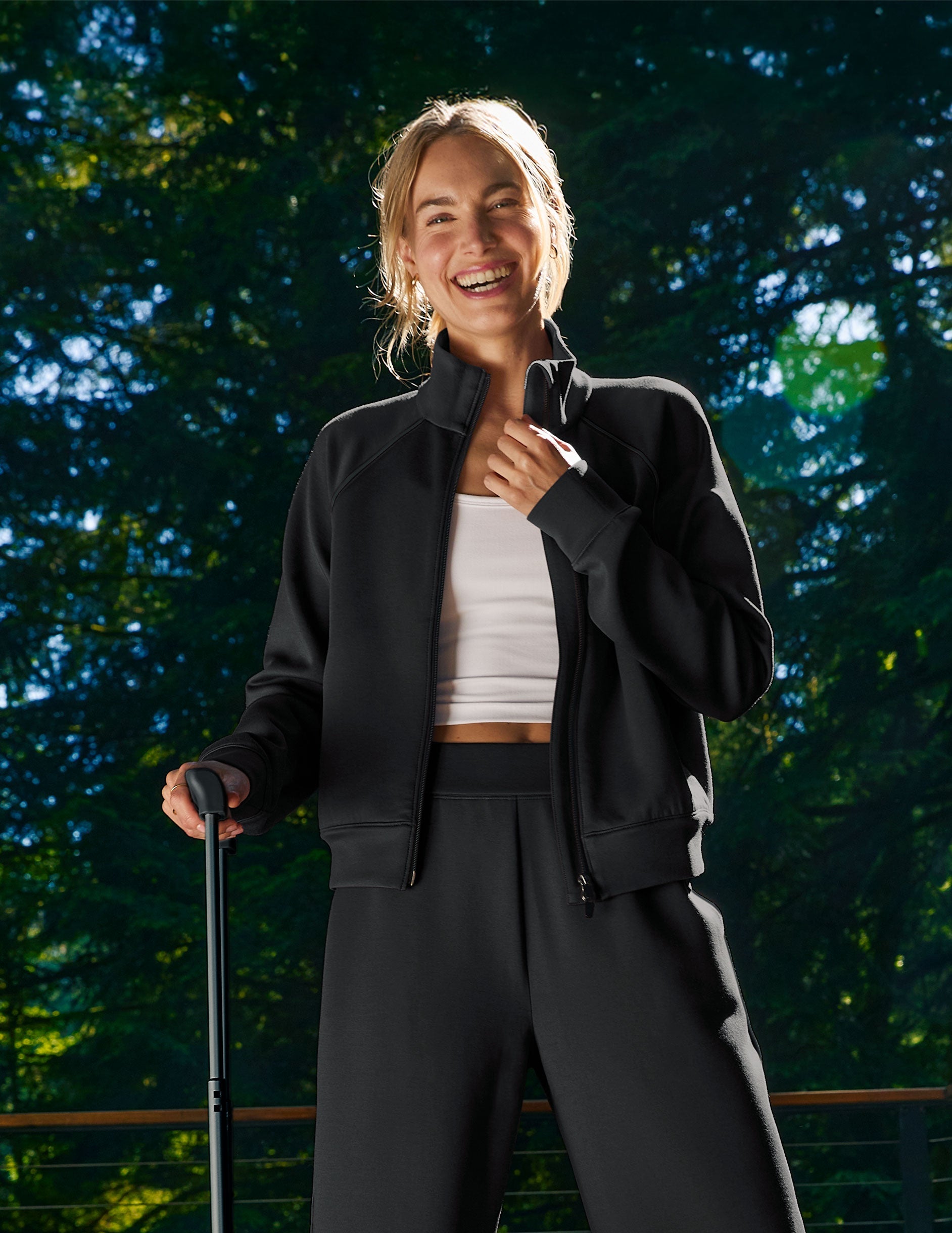 Woman in black jacket and pants standing outdoors with a blurred green background.