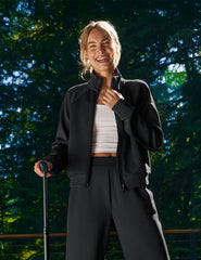 Woman in black jacket and pants standing outdoors with a blurred green background.