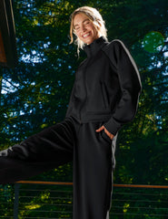 Woman in black jacket and pants standing outdoors with a blurred green background.