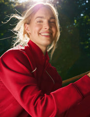 woman wearing a red zip-up jacket with white piping against a nature background.