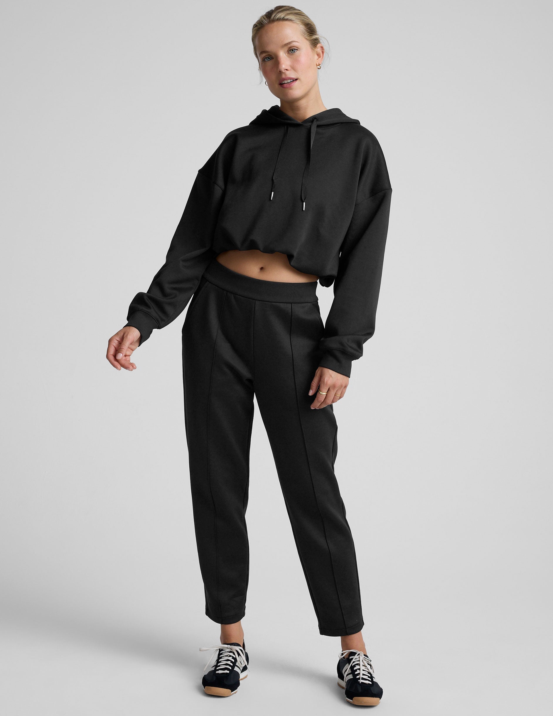 woman wearing a black cropped hoodie and pants on a plain background.