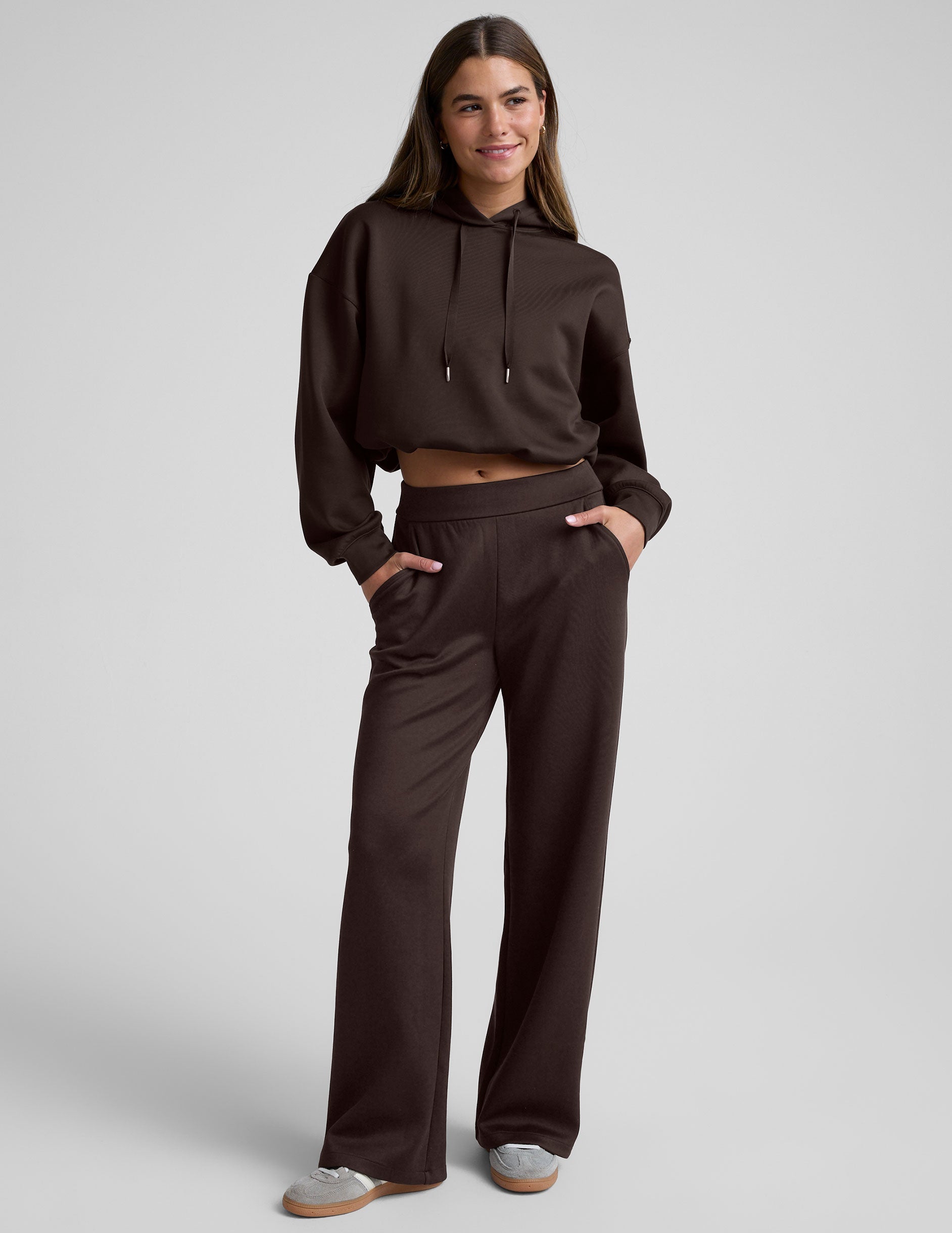 woman wearing a brown cropped hoodie and pants against a plain background.