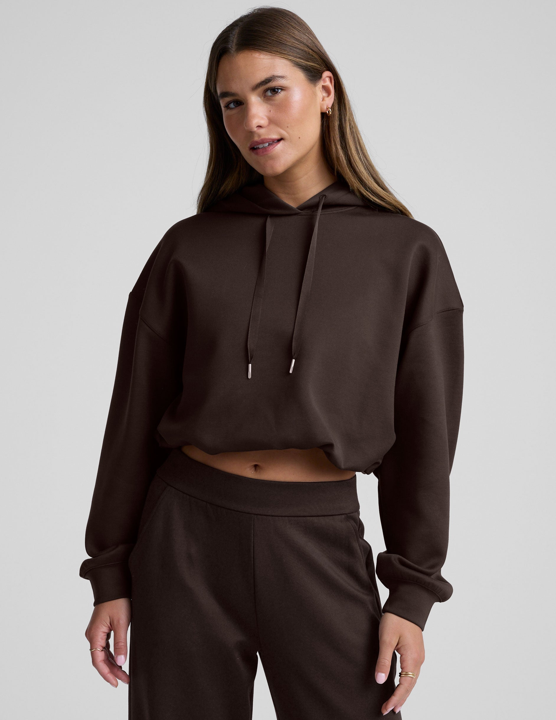 woman wearing a brown cropped hoodie and pants against a plain background.