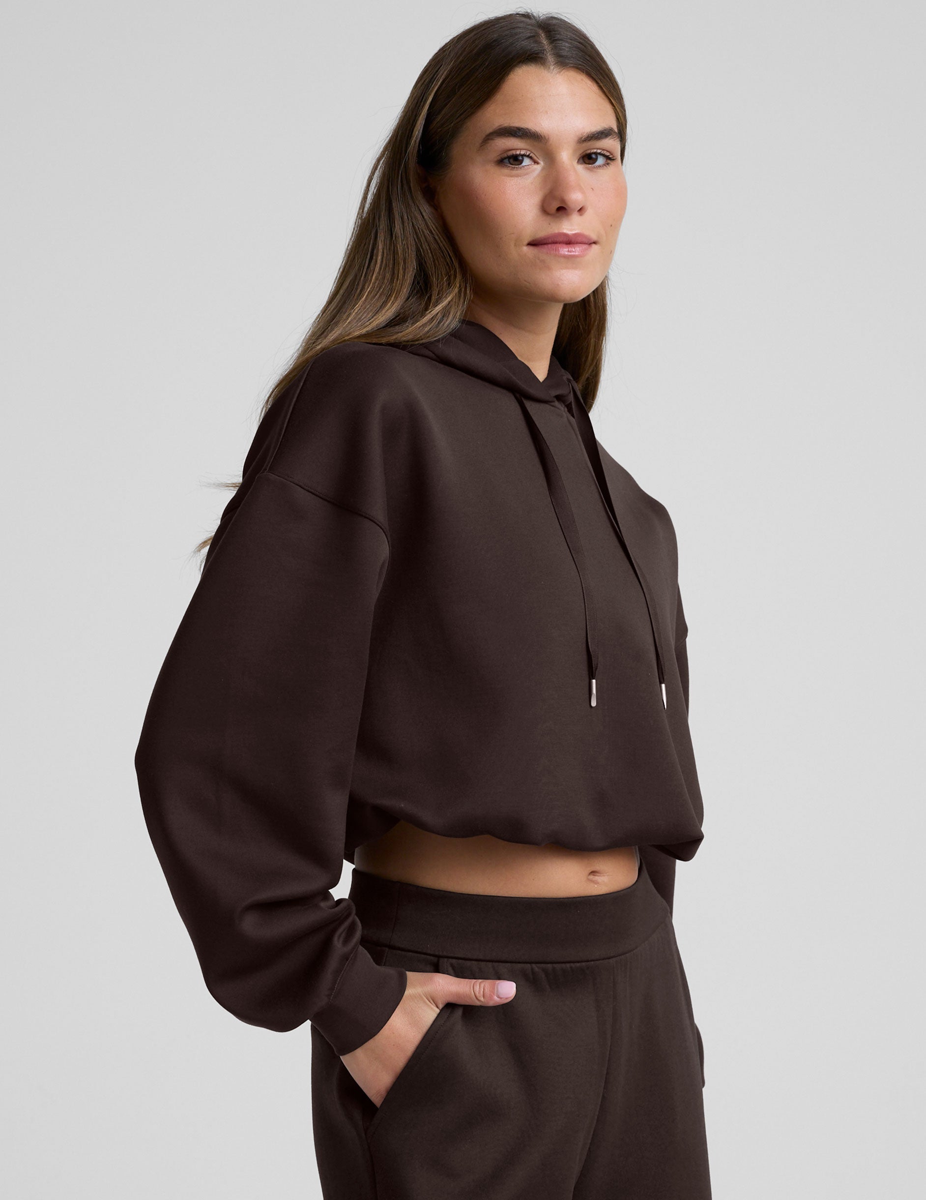 woman wearing a brown cropped hoodie and pants against a plain background.
