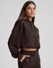 woman wearing a brown cropped hoodie and pants against a plain background.