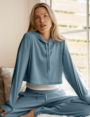 woman wearing a blue ribbed cropped hoodie and pants sitting in a room with a window in the background.