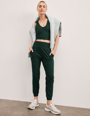 woman wearing a green cropped tank and joggers with a gray jacket against a plain background.