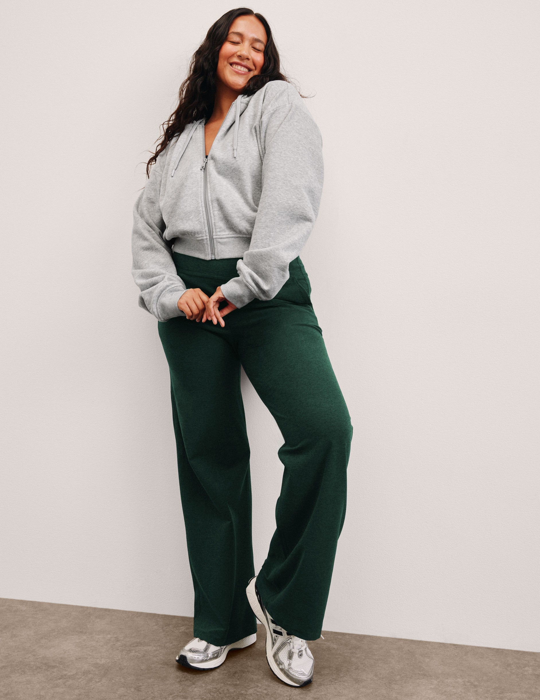 woman wearing a gray zip-up hoodie and green pants against a plain background.