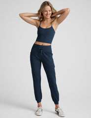 model wearing a navy blue cropped tank and navy blue high-waisted midi length joggers with a drawcord at waistband and pockets athletic outfit on a plain background.