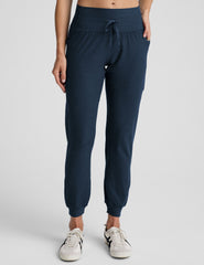 model wearing navy blue high-waisted midi length joggers with a drawcord at waistband and pockets athletic outfit on a plain background.