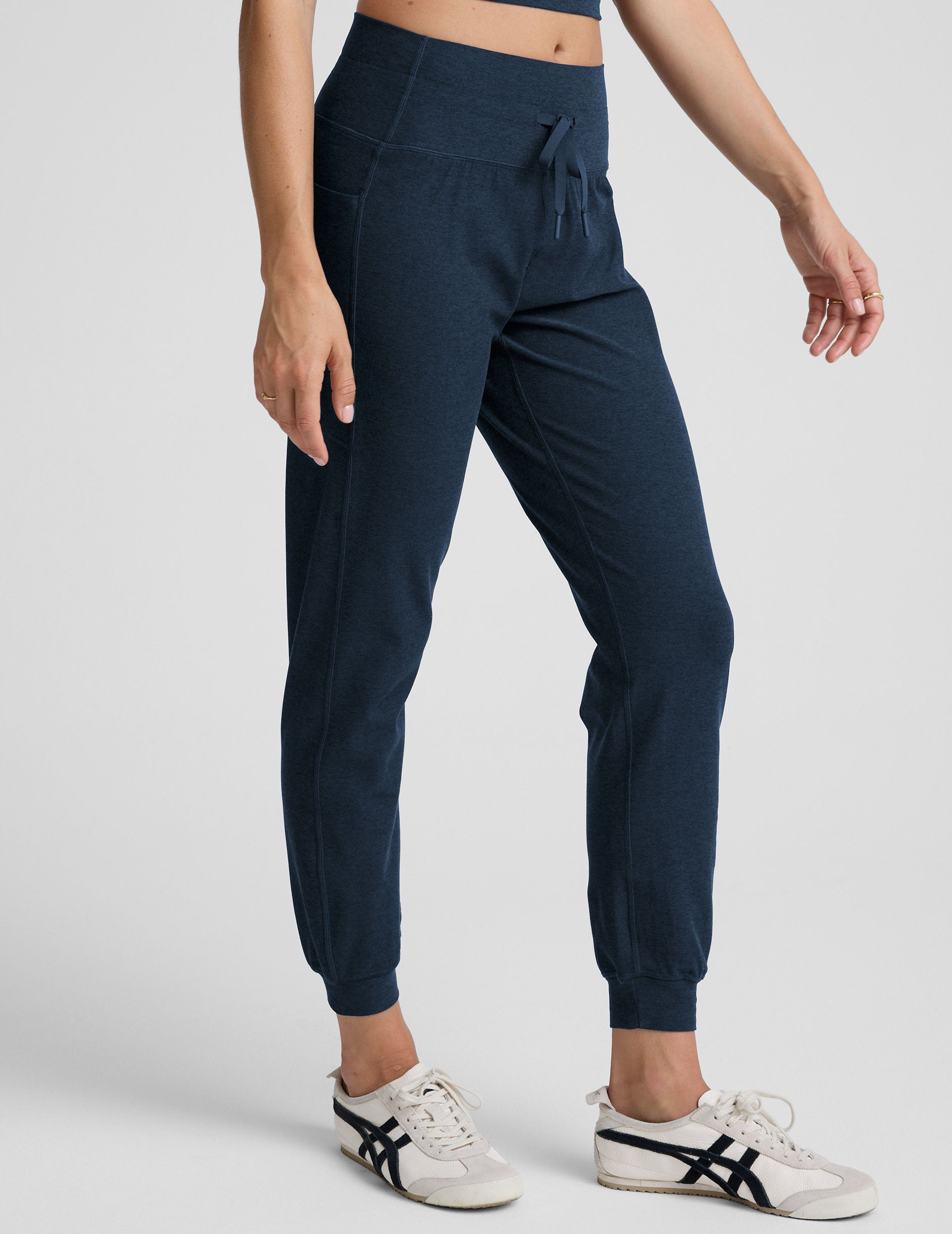 model wearing navy blue high-waisted midi length joggers with a drawcord at waistband and pockets athletic outfit on a plain background.