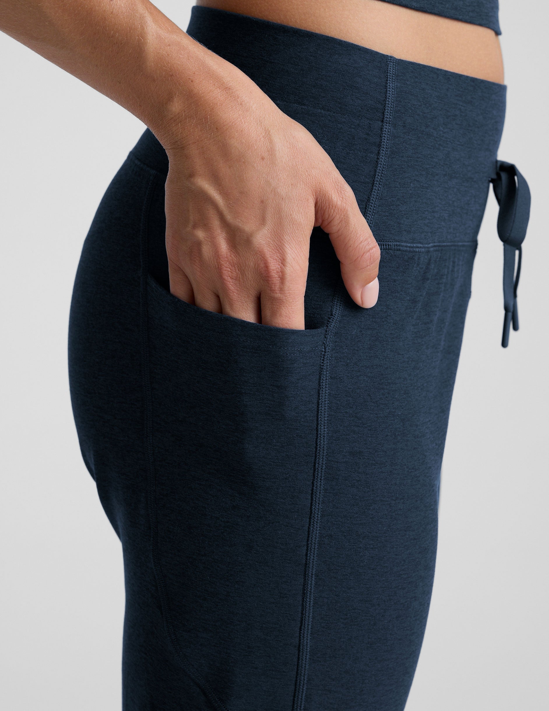 model wearing navy blue high-waisted midi length joggers with a drawcord at waistband and pockets athletic outfit on a plain background.