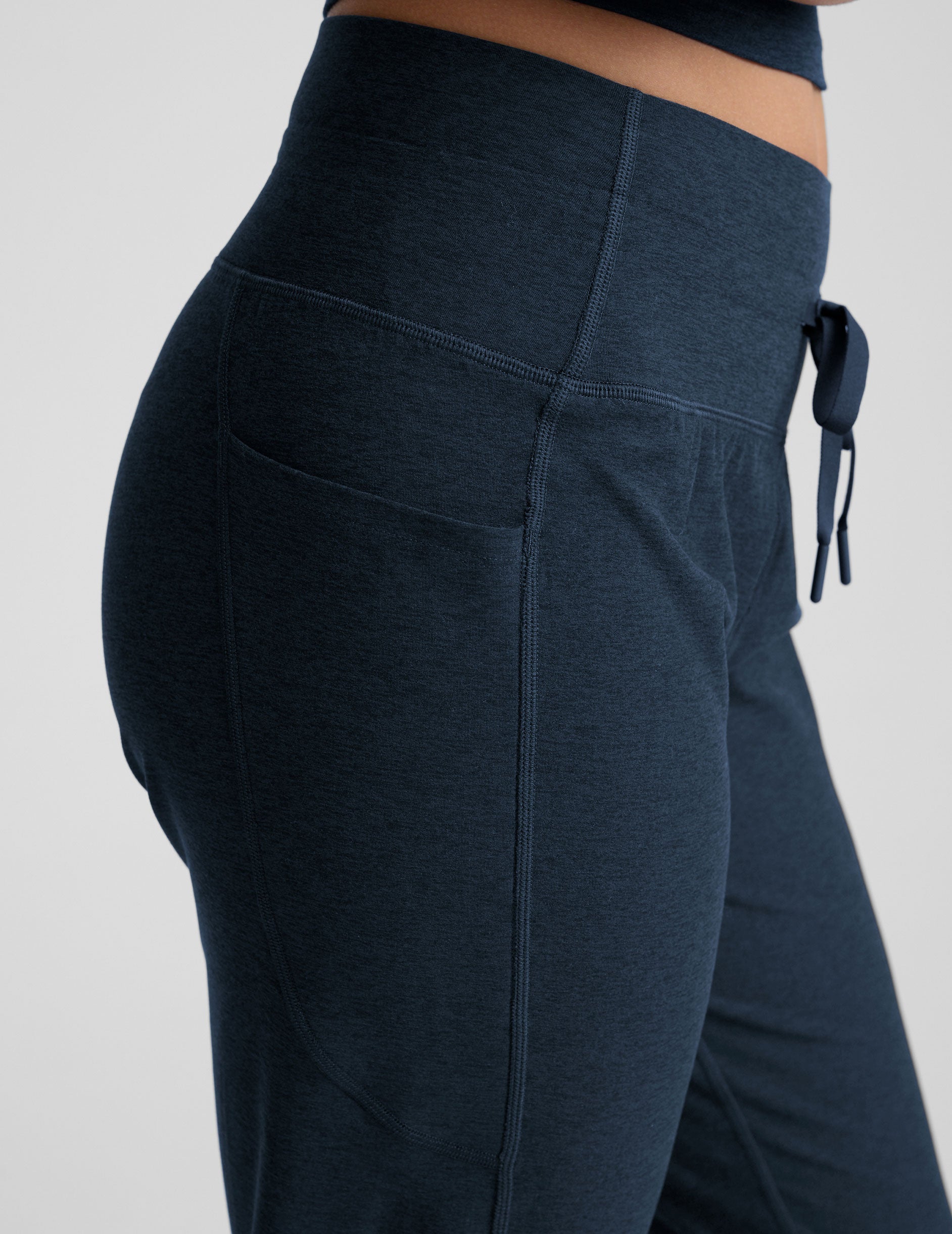 model wearing navy blue high-waisted midi length joggers with a drawcord at waistband and pockets athletic outfit on a plain background.