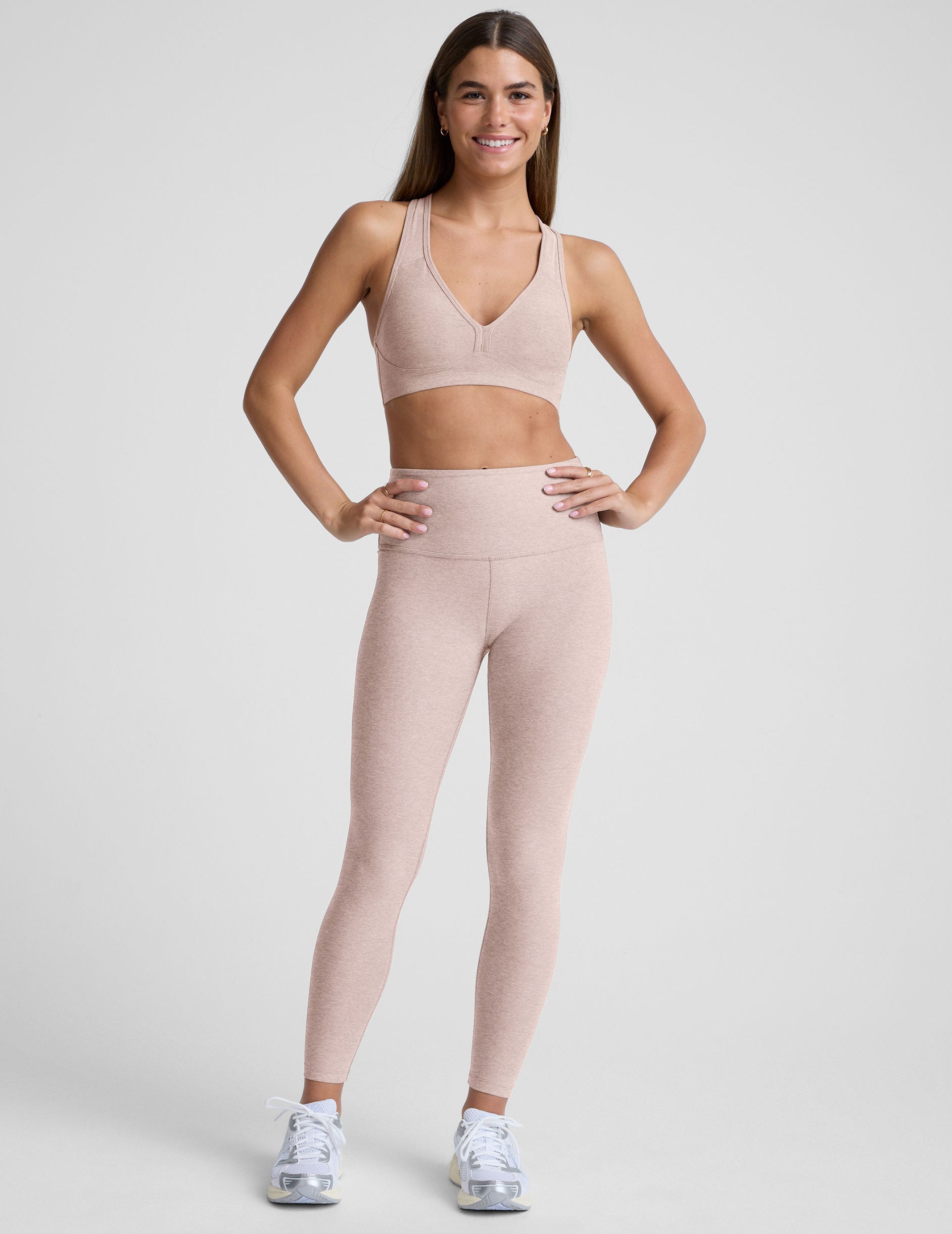 model is wearing a light pink sports bra and high-waisted midi length leggings on a light gray background.