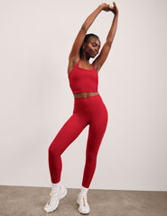 woman wearing a red slim racerback cropped tank top and high-waisted midi length leggings against a plain background.