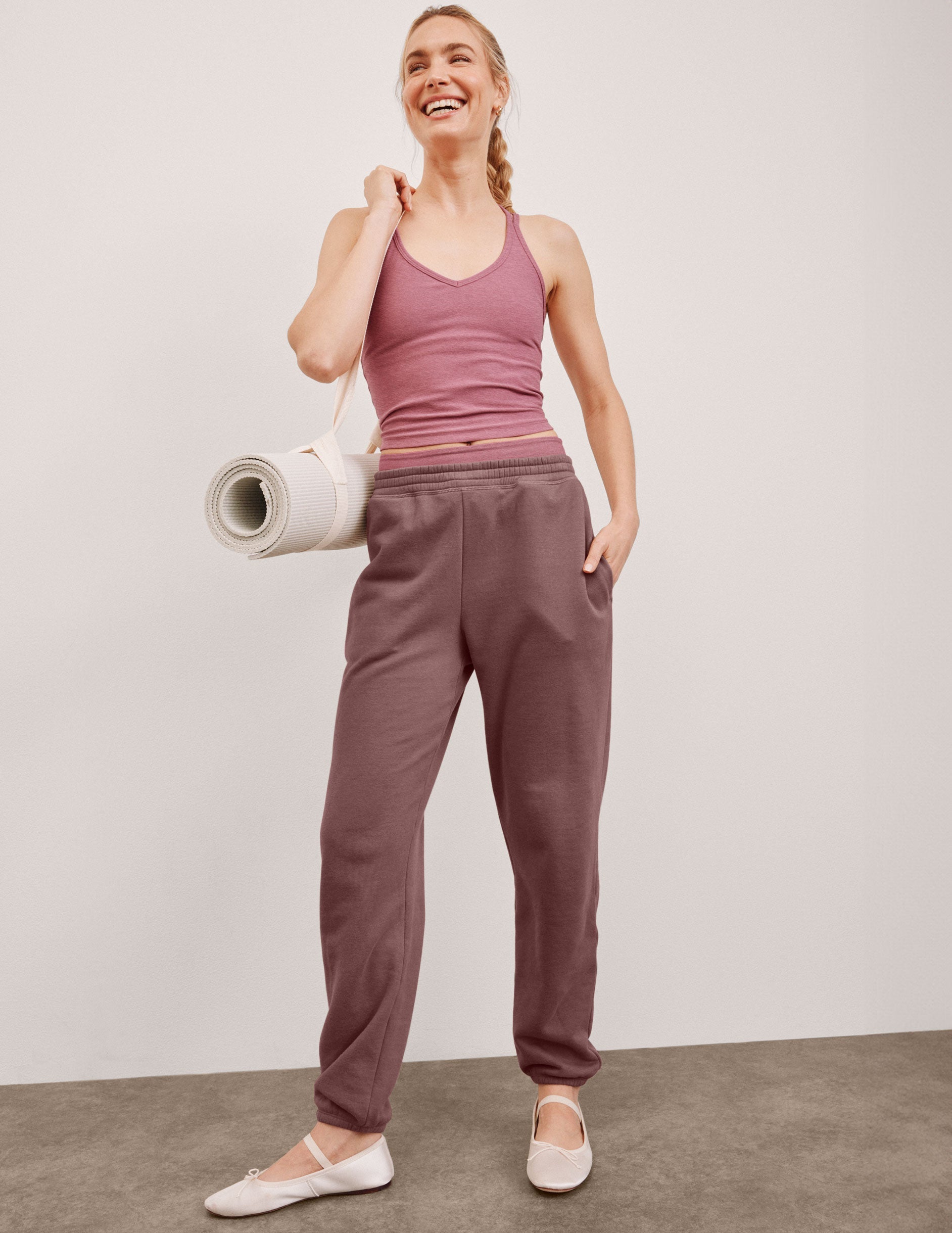 model in pink tank top and brown sweatpants holding a yoga mat against a plain background.