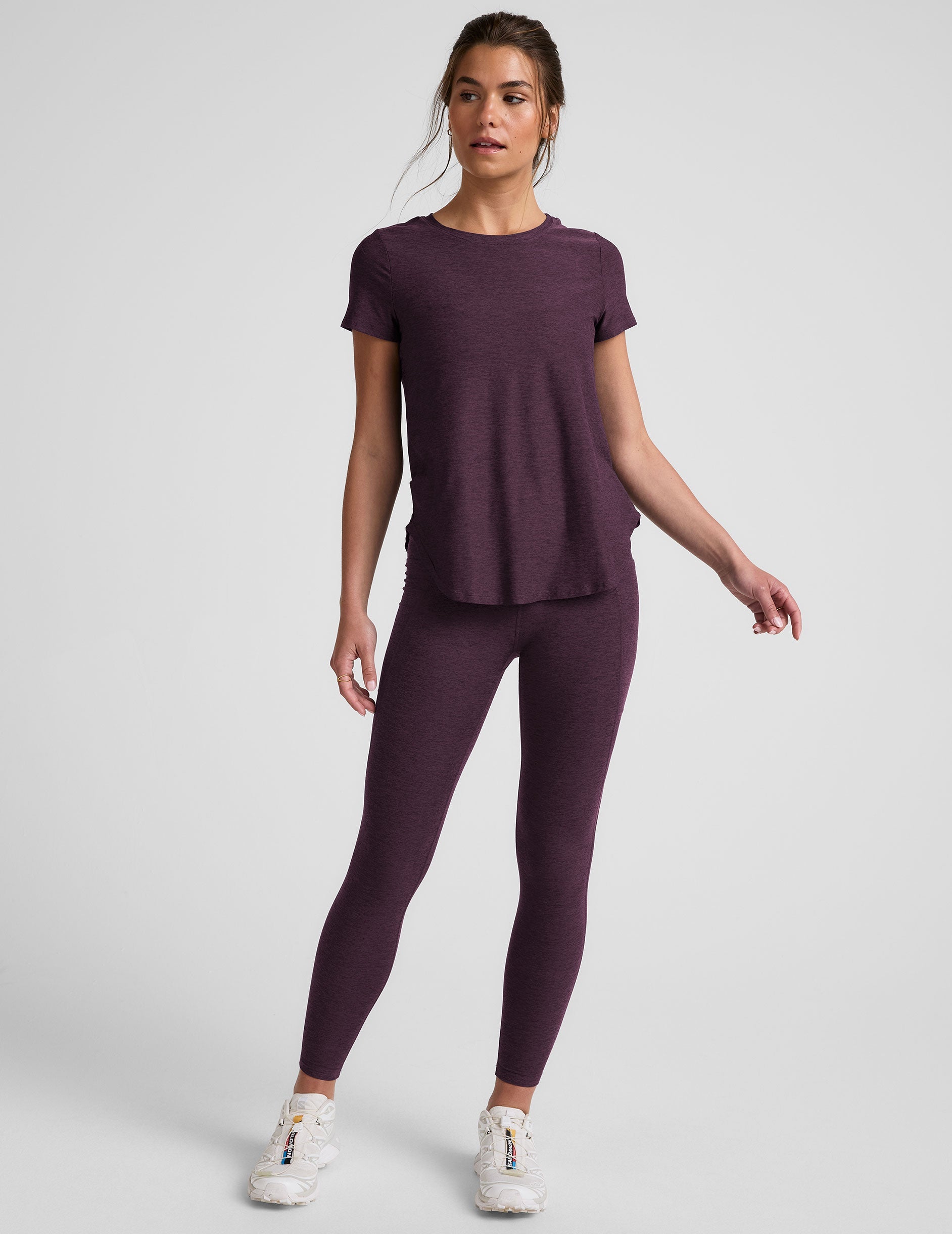 model wearing a purple t-shirt and purple high-waisted midi length leggings with pockets on a light gray background.