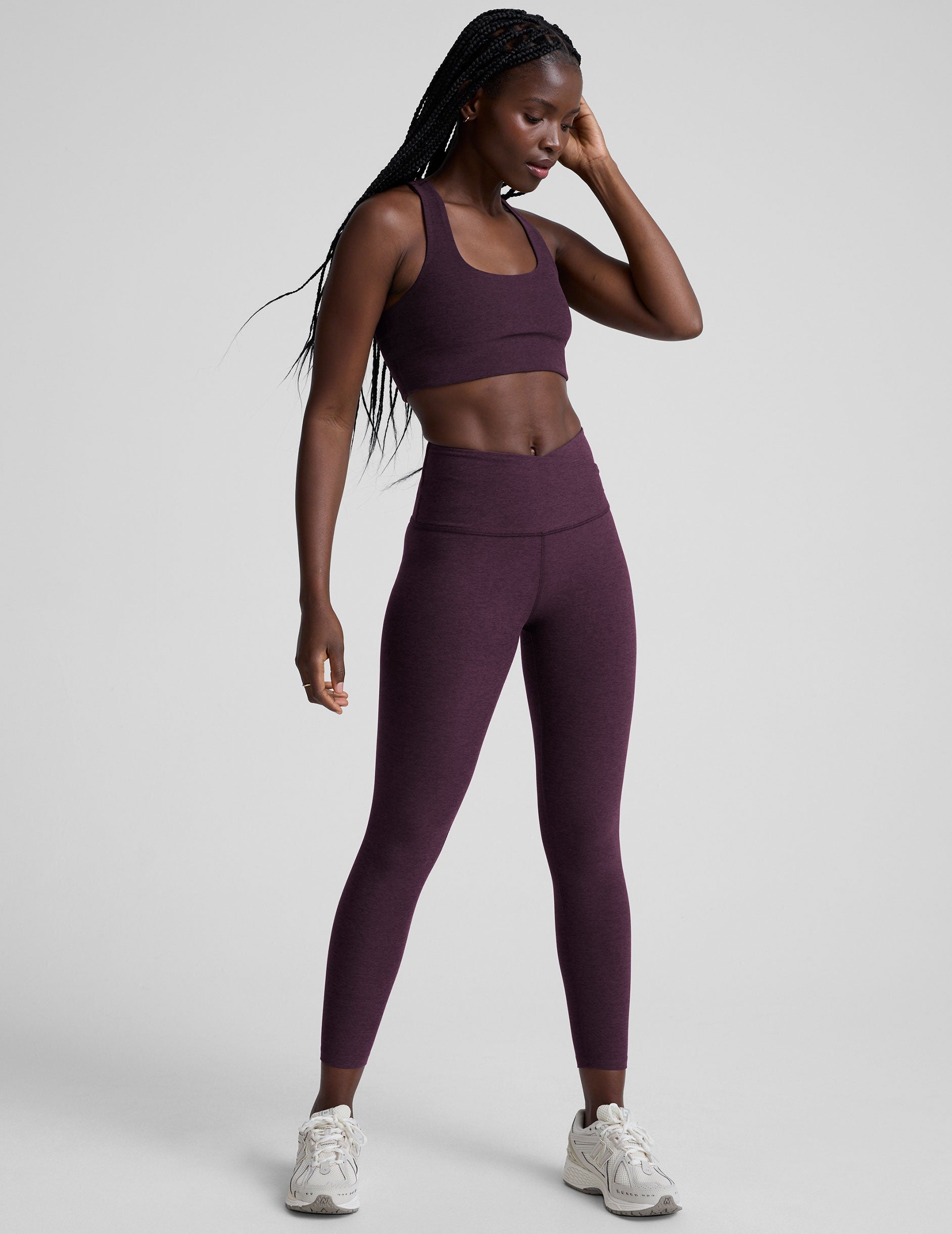 model wearing a purple bra with purple high-waisted midi length leggings with a front crossover detail on the front waistband on a plain background.