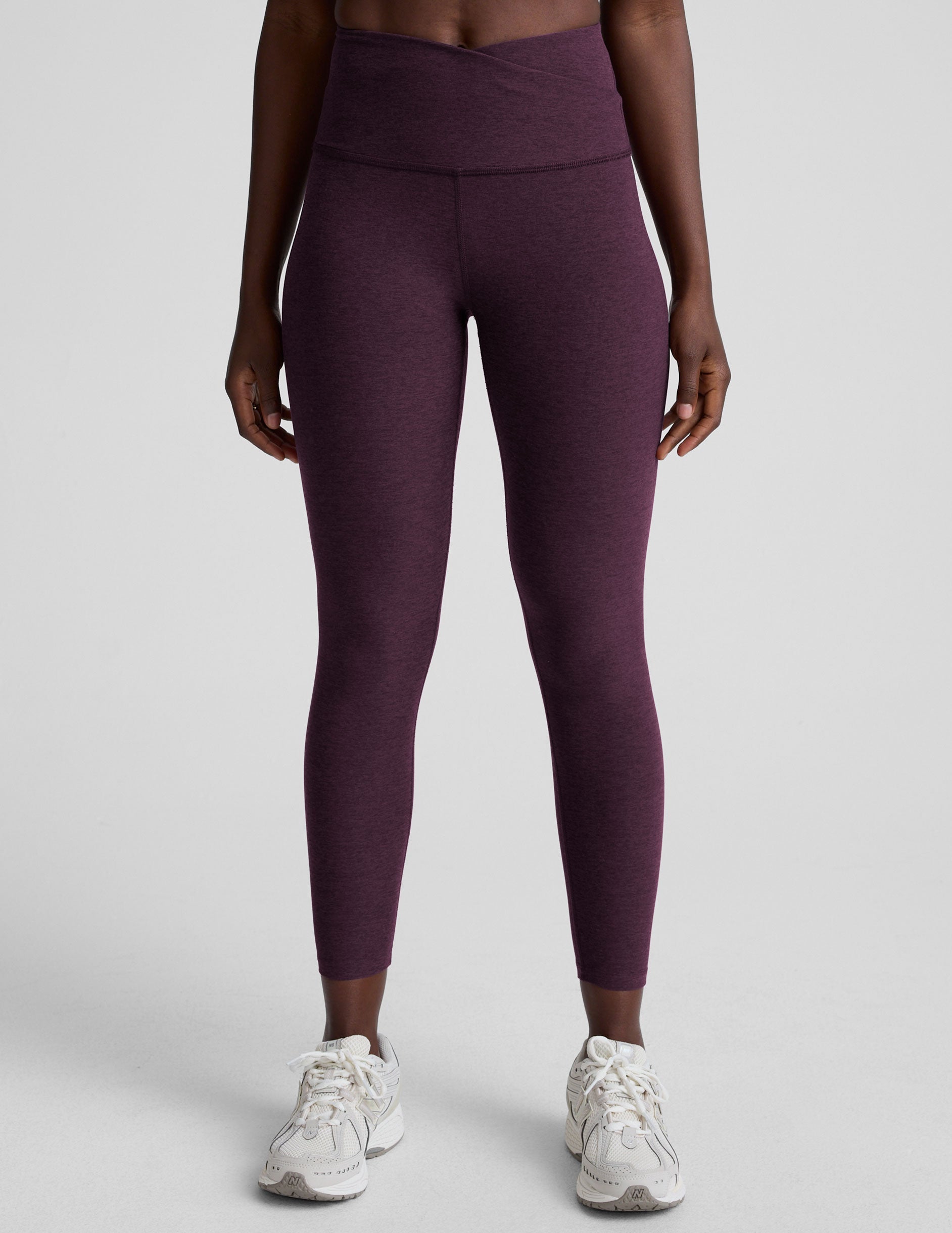 model wearing purple high-waisted midi length leggings with a front crossover detail on the waistband on a plain background.