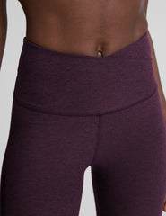 model wearing purple high-waisted midi length leggings with a front crossover detail on the waistband on a plain background.