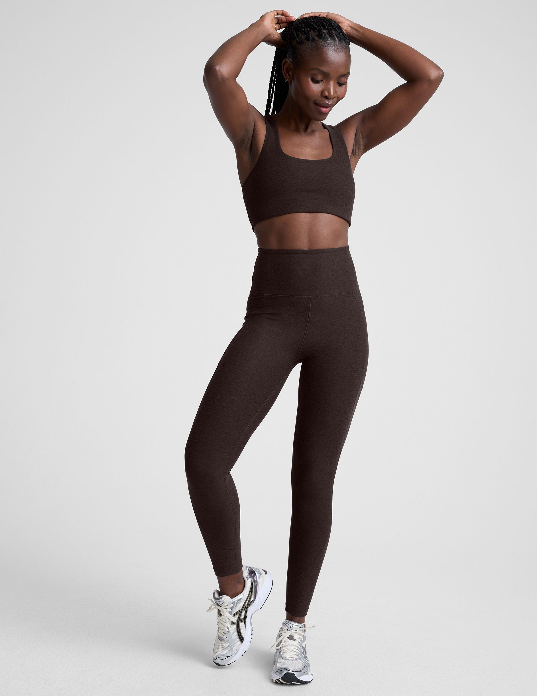 woman wearing brown high-waisted midi length leggings with pockets on a plain background.