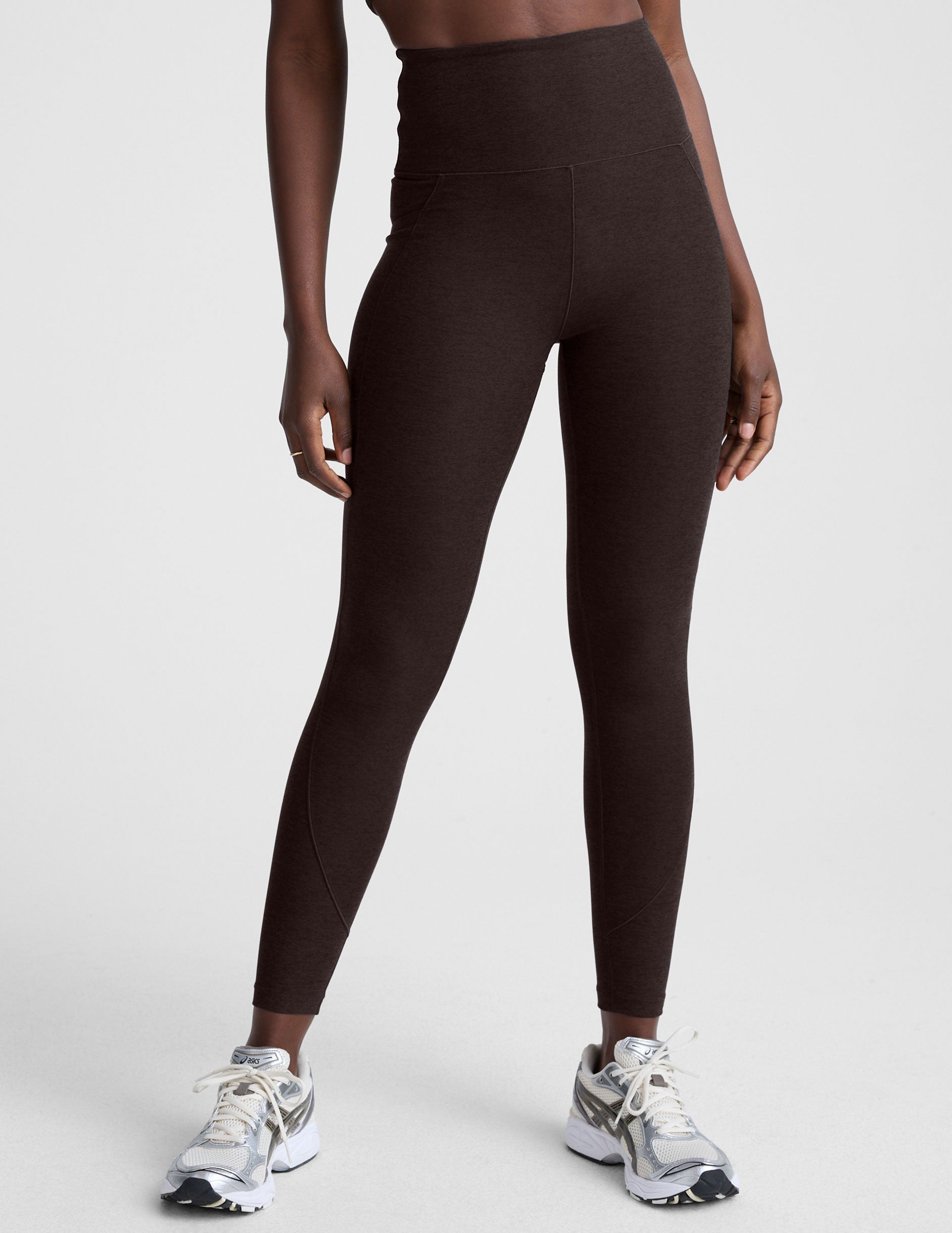 woman wearing brown high-waisted midi length leggings with pockets on a plain background.