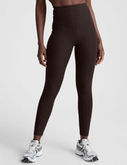 woman wearing brown high-waisted midi length leggings with pockets on a plain background.