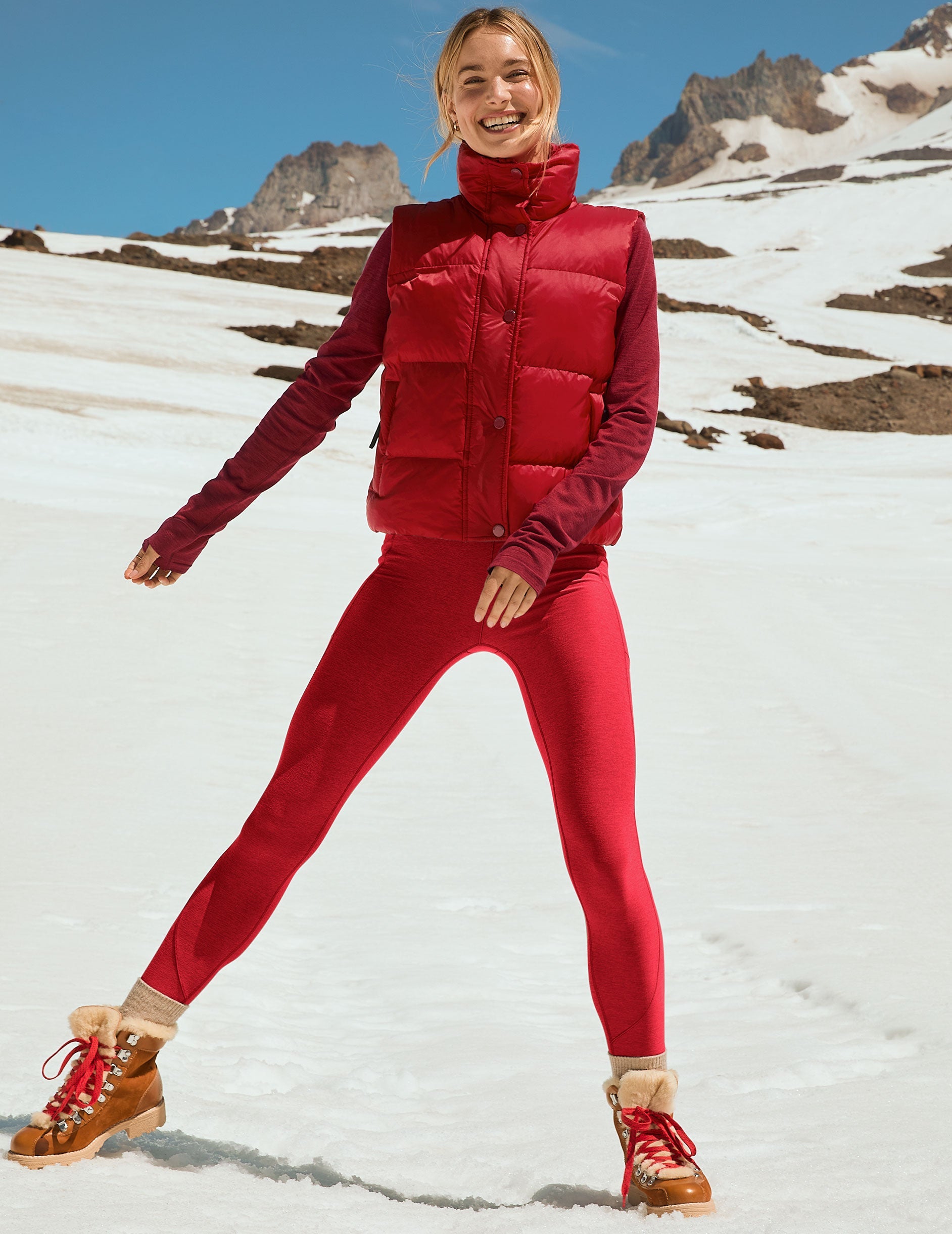 woman wearing a red puffer vest and leggings with pockets standing on a snowy mountain.