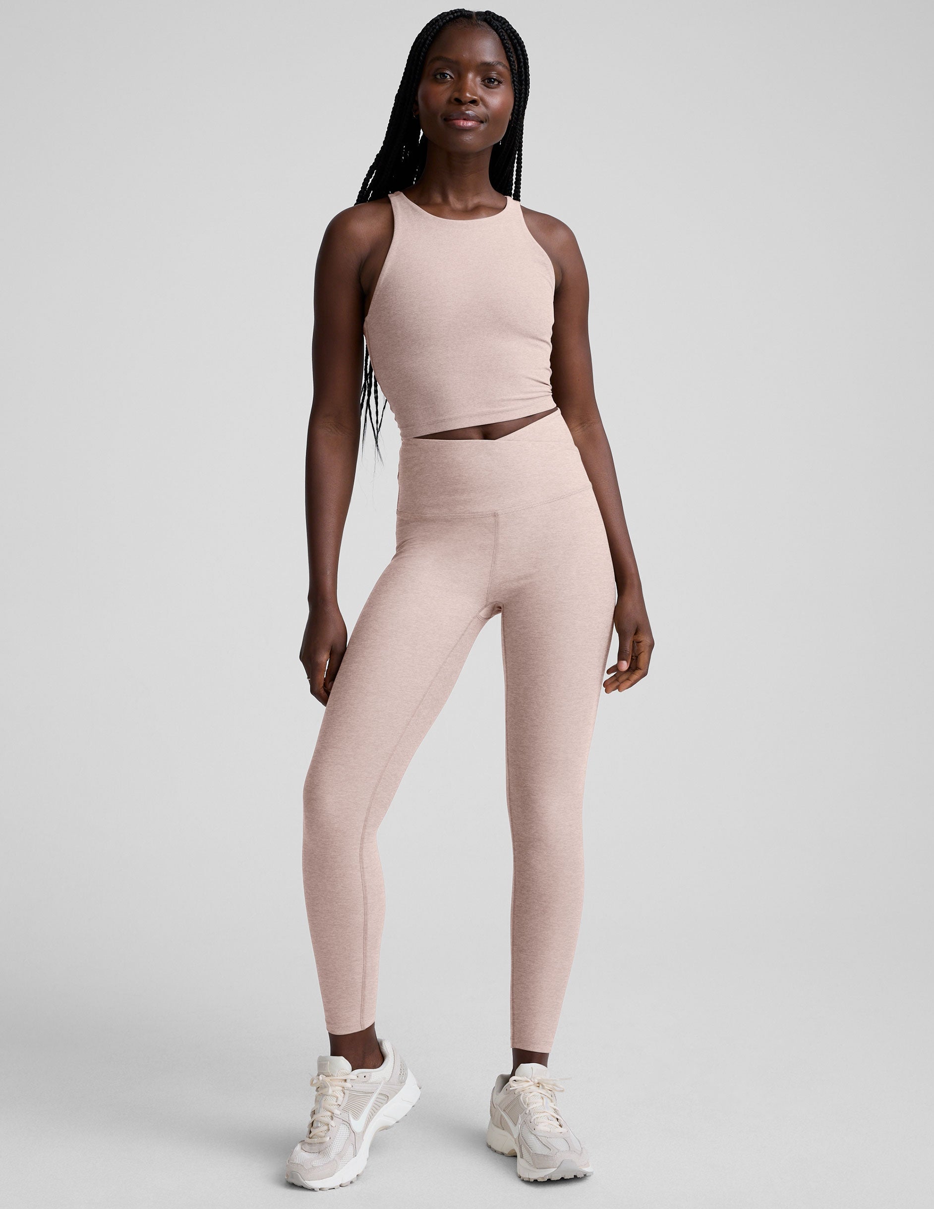 model wearing a beige high neck racerback cropped tank top and beige high-waisted midi length leggings against a plain background.