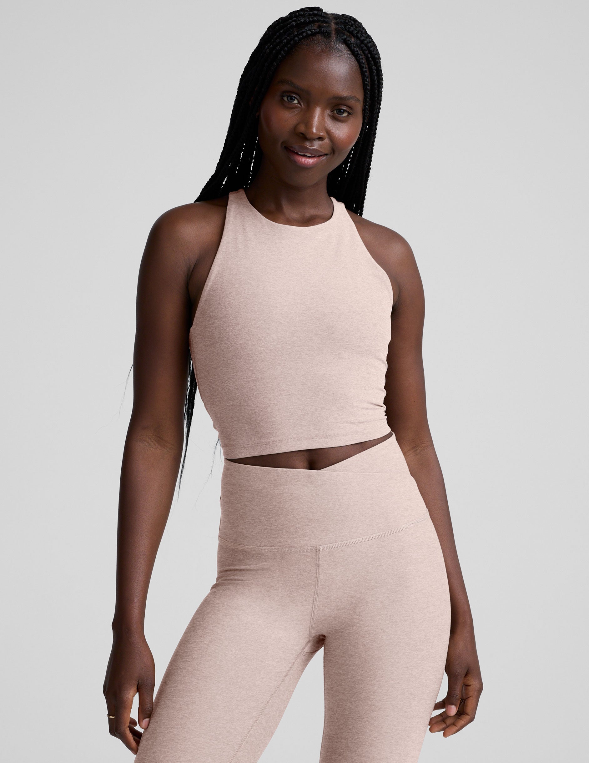 model wearing a beige high neck racerback cropped tank top and beige high-waisted midi length leggings against a plain background.