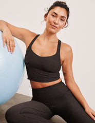 Woman in brown v-neck cropped racerback tank and brown leggings sitting next to a blue exercise ball against a plain background.