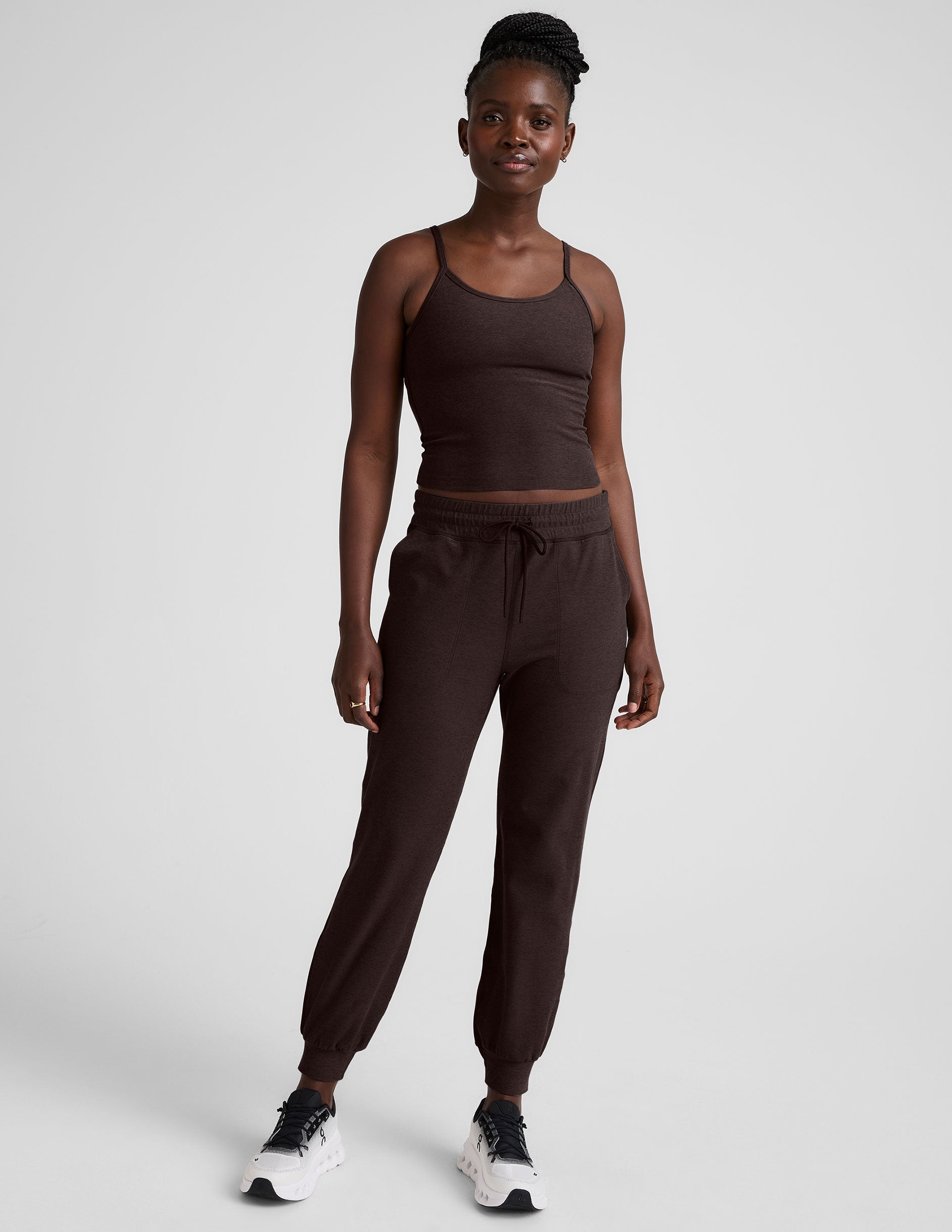 Woman wearing a brown midway length tank top with adjustable straps and brown jogger pants against a plain background.