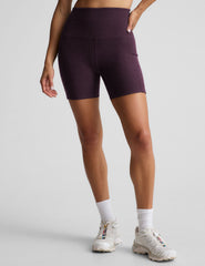 model is wearing a purple sports bra and purple 5" biker shorts with pockets against a plain background. 