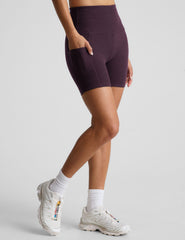 model is wearing a purple sports bra and purple 5" biker shorts with pockets against a plain background. 