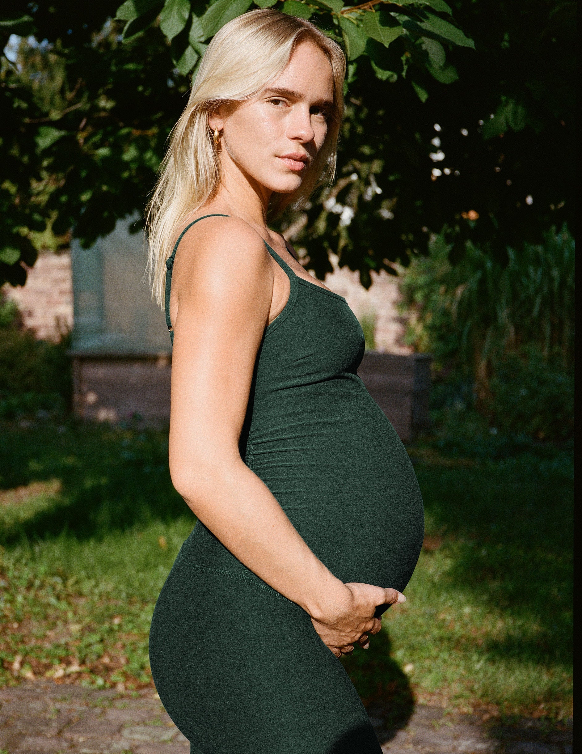 pregnant woman green midi length jumpsuit standing outdoors with trees in the background.