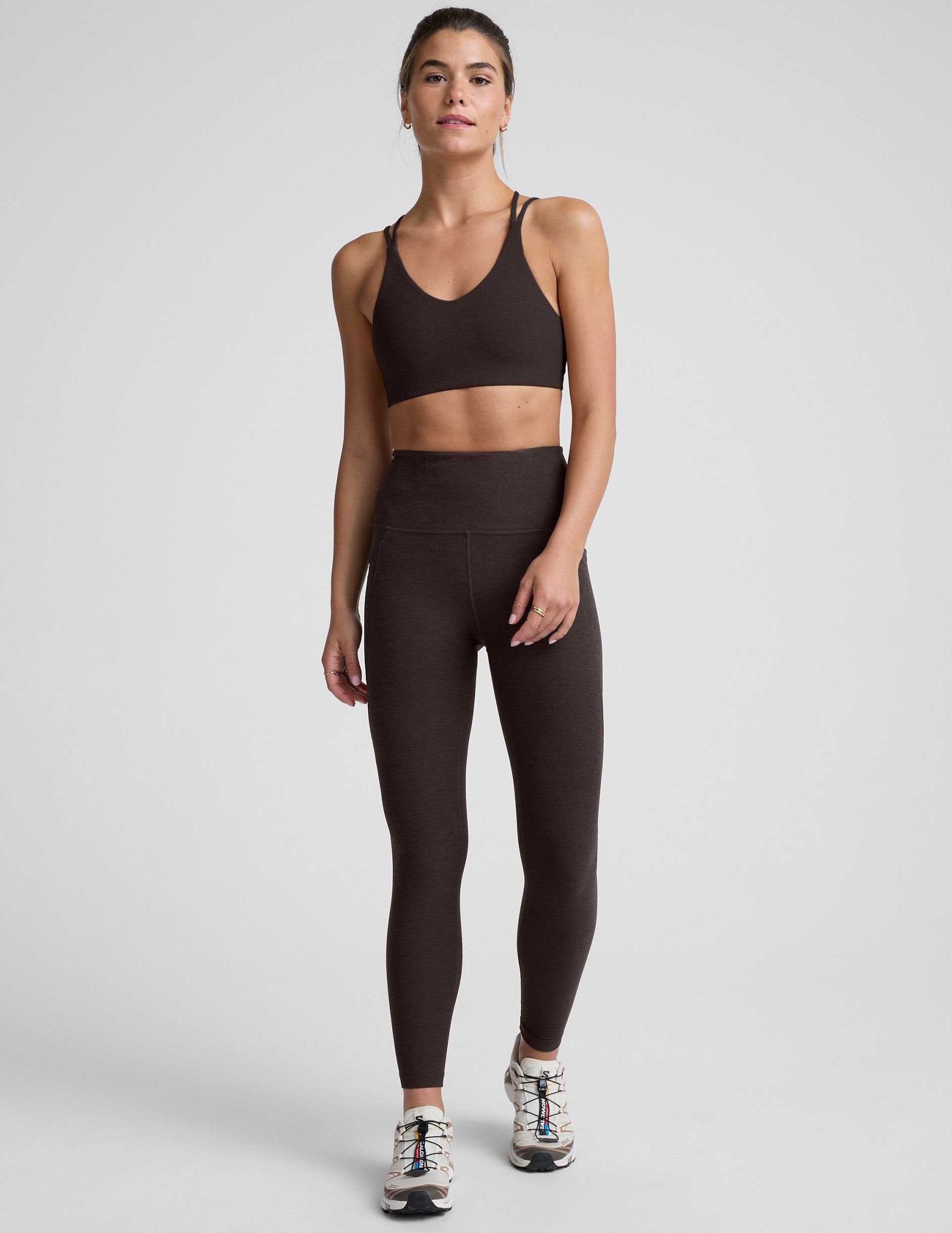 woman wearing a brown sports bra with crossover back straps and leggings on a plain background.