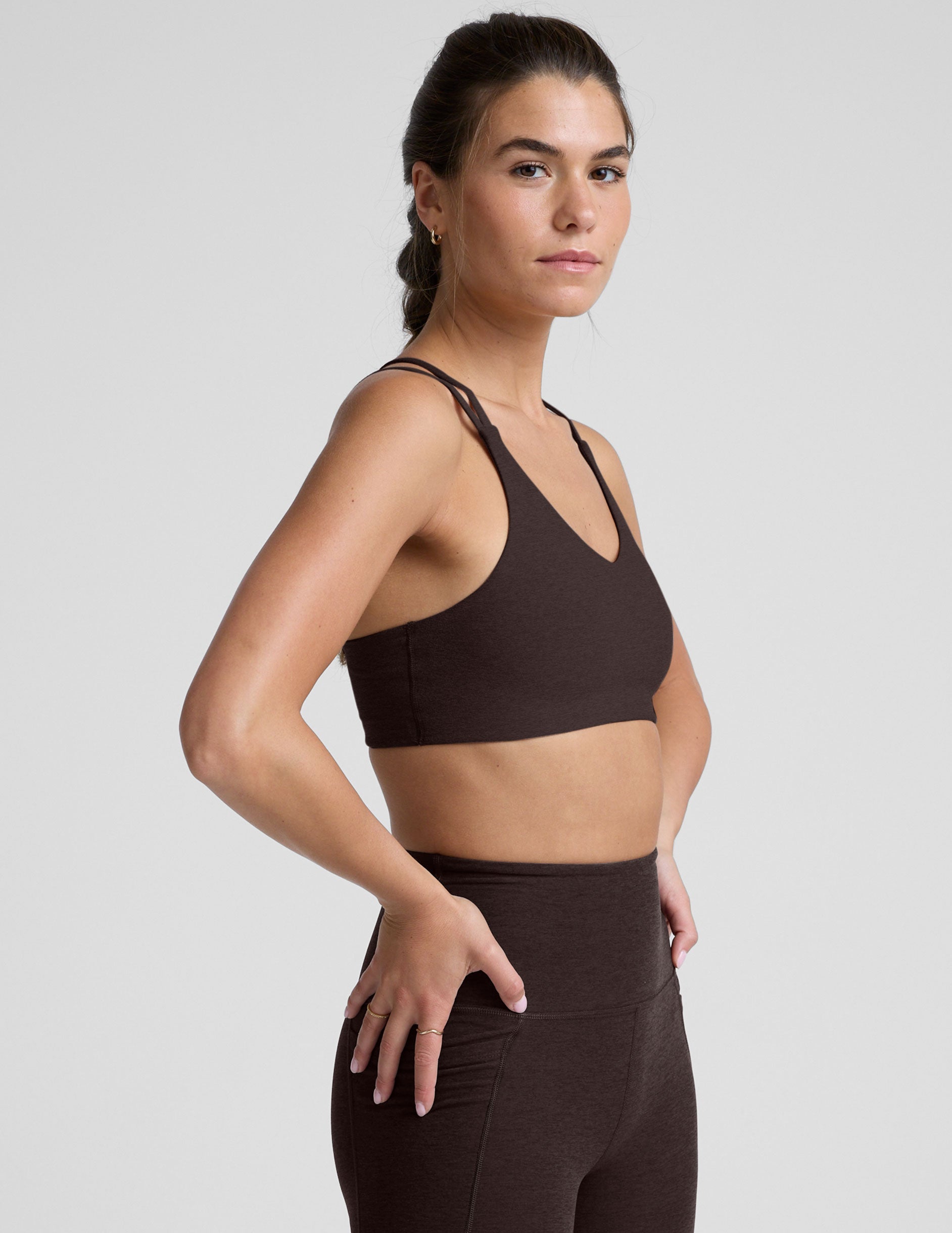 woman wearing a brown sports bra with crossover back straps and leggings on a plain background.