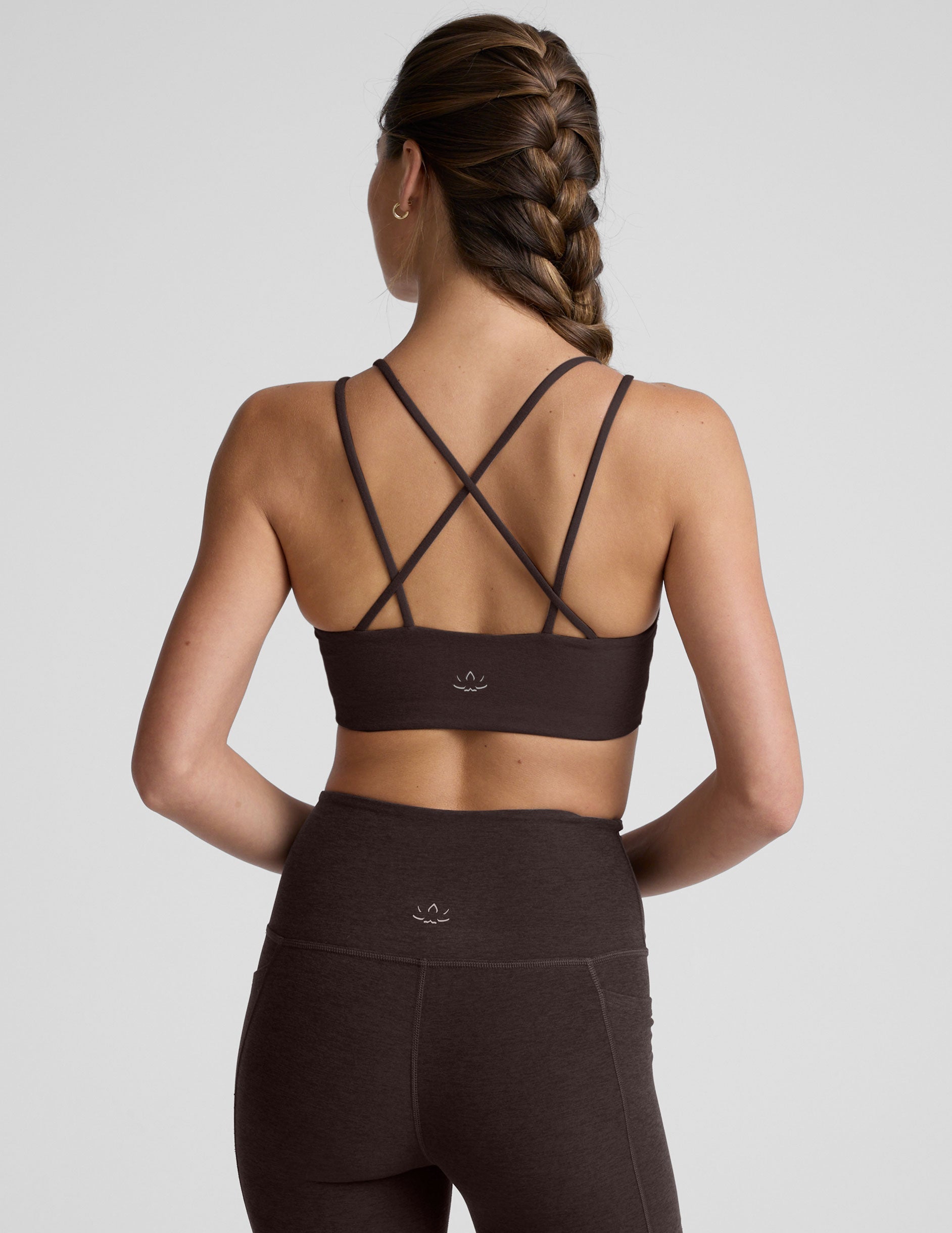 woman wearing a brown sports bra with crossover back straps and leggings on a plain background.